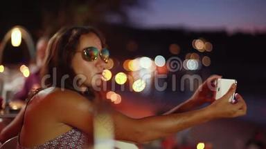 年轻漂亮的黑发女人，戴着太阳镜，摆着假脸，用智能手机和闪光灯自拍。